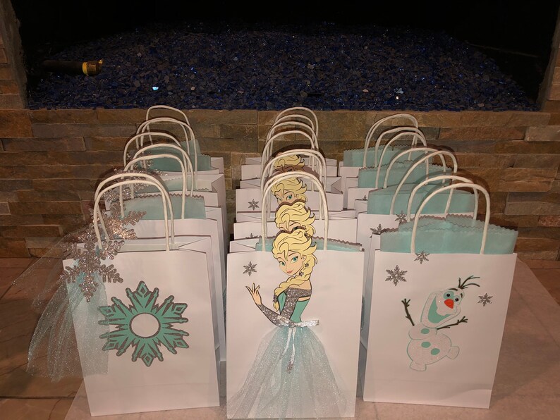 Może przedstawiać: Zbi&oacute;r białych papierowych toreb na prezenty z niebieskimi i srebrnymi wzorami śnieżynek. Niekt&oacute;re torby przedstawiają kresk&oacute;wkową postać Elsy z Krainy Lodu, a inne Olafa.