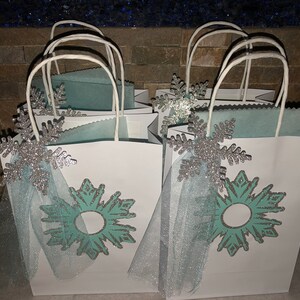 Może przedstawiać: Białe torby prezentowe z niebieskimi i srebrnymi wzorami płatk&oacute;w śniegu i kokardkami z tiulu. Torby są ozdobione turkusowym płatkiem śniegu z srebrnym błyszczącym obramowaniem.
