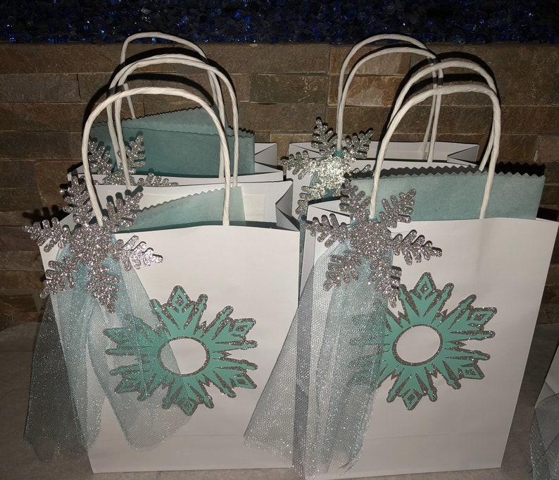 Może przedstawiać: Białe torby prezentowe z niebieskimi i srebrnymi wzorami płatk&oacute;w śniegu i kokardkami z tiulu. Torby są ozdobione turkusowym płatkiem śniegu z srebrnym błyszczącym obramowaniem.