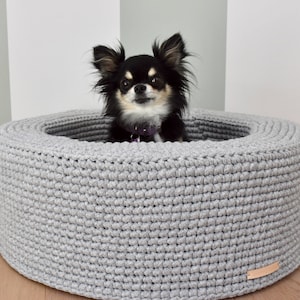 Pode incluir: Uma cama de crochet cinza para cães com um chihuahua preto e castanho sentado dentro. A cama é redonda e tem uma borda elevada.