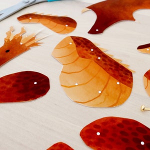 Puede incluir: Recortes de papel con forma de drag&oacute;n en tonos naranja y marr&oacute;n, con detalles de puntos blancos. Las piezas est&aacute;n dispuestas sobre una superficie de madera clara, con unas tijeras y un cierre dorado.