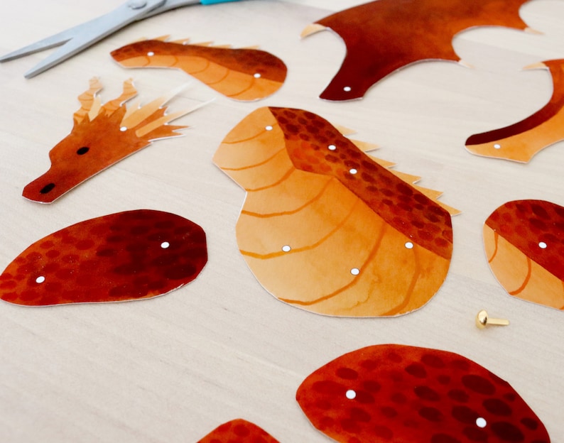 Puede incluir: Recortes de papel con forma de drag&oacute;n en tonos naranja y marr&oacute;n, con detalles de puntos blancos. Las piezas est&aacute;n dispuestas sobre una superficie de madera clara, con unas tijeras y un cierre dorado.