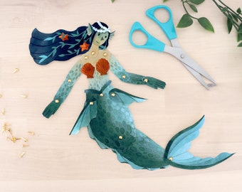 Puede incluir: Muñeca de sirena de papel con articulaciones, cola verde azulado, sujetador de concha y cabello azul oscuro con detalles de estrellas naranjas. Incluye unas tijeras con mango azul y pequeños brads dorados.