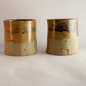 May include: Two cylindrical ceramic cups featuring a tri-tone glaze. The base is a speckled matte beige, transitioning into a smooth mustard yellow middle section, and finished with a dark brown, glossy rim. Each cup has a small stamped mark.