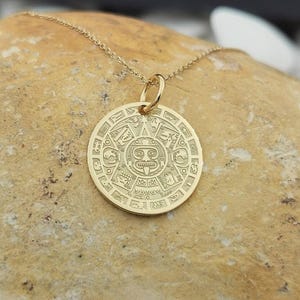 May include: A gold-toned circular pendant necklace featuring an intricate embossed design of a central face surrounded by geometric patterns and symbols, suspended from a delicate matching gold-toned chain link necklace.