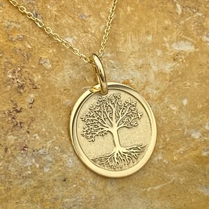 May include: A gold-colored pendant necklace featuring a circular charm with a detailed tree design. The tree has intricate branches and roots, set against a textured background. The necklace chain is also gold-colored.