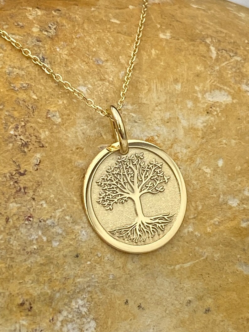 May include: A gold-colored pendant necklace featuring a circular charm with a detailed tree design. The tree has intricate branches and roots, set against a textured background. The necklace chain is also gold-colored.