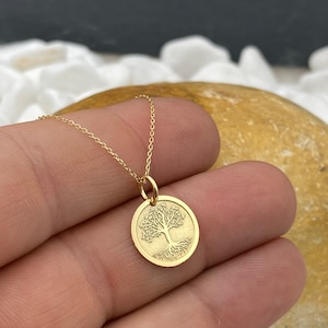 May include: A gold-colored necklace featuring a circular pendant with a detailed tree design. The tree has a trunk, branches, and roots. The necklace chain is delicate and thin. The pendant is approximately 0.75 inches in diameter.