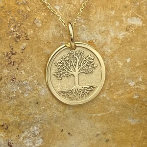 May include: A gold-colored pendant necklace featuring a circular charm with a detailed tree of life design. The tree has intricate branches and roots. The necklace chain is delicate and also gold-colored.