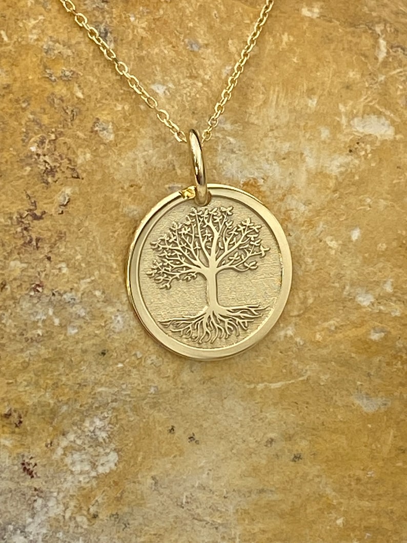 May include: A gold-colored pendant necklace featuring a circular charm with a detailed tree of life design. The tree has intricate branches and roots. The necklace chain is delicate and also gold-colored.