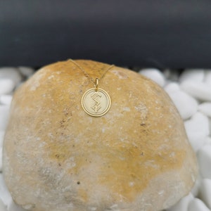 May include: Gold pendant necklace with a circular charm featuring a geometric design. The necklace is displayed on a light brown rock.