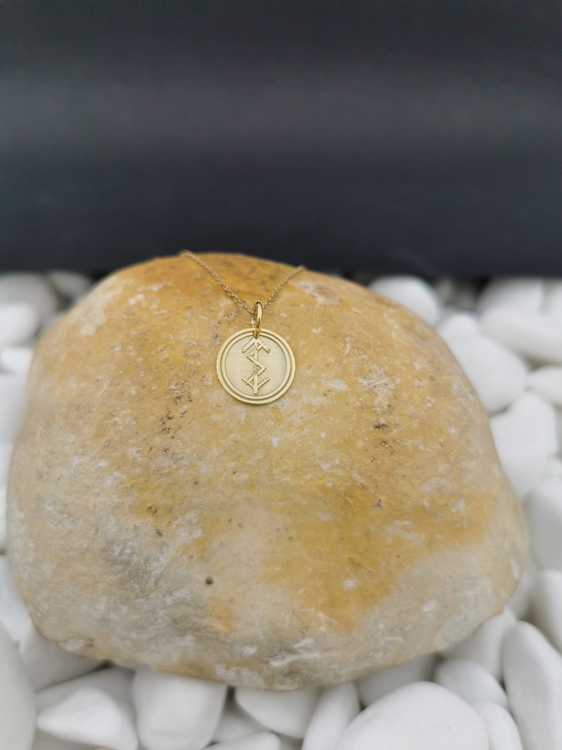 May include: Gold pendant necklace with a circular charm featuring a geometric design. The necklace is displayed on a light brown rock.