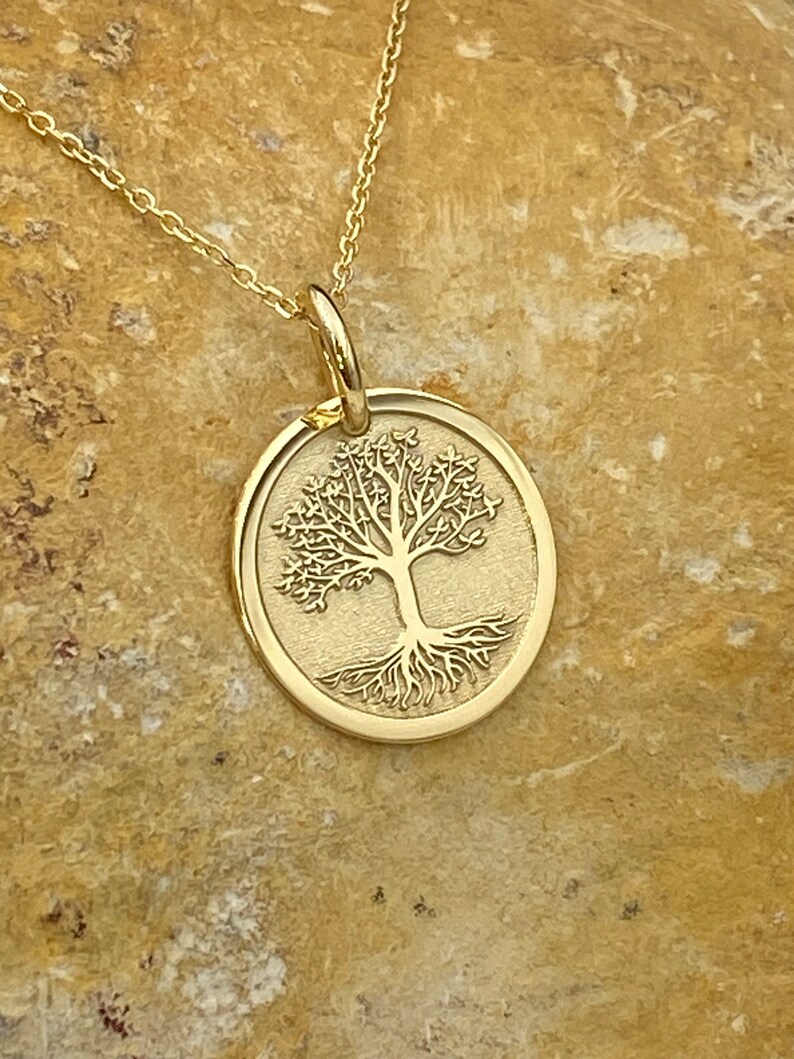 May include: A gold-colored pendant necklace featuring a detailed tree design within a circular frame. The tree has intricate branches and roots. The necklace chain is also gold and is displayed on a textured, tan surface.