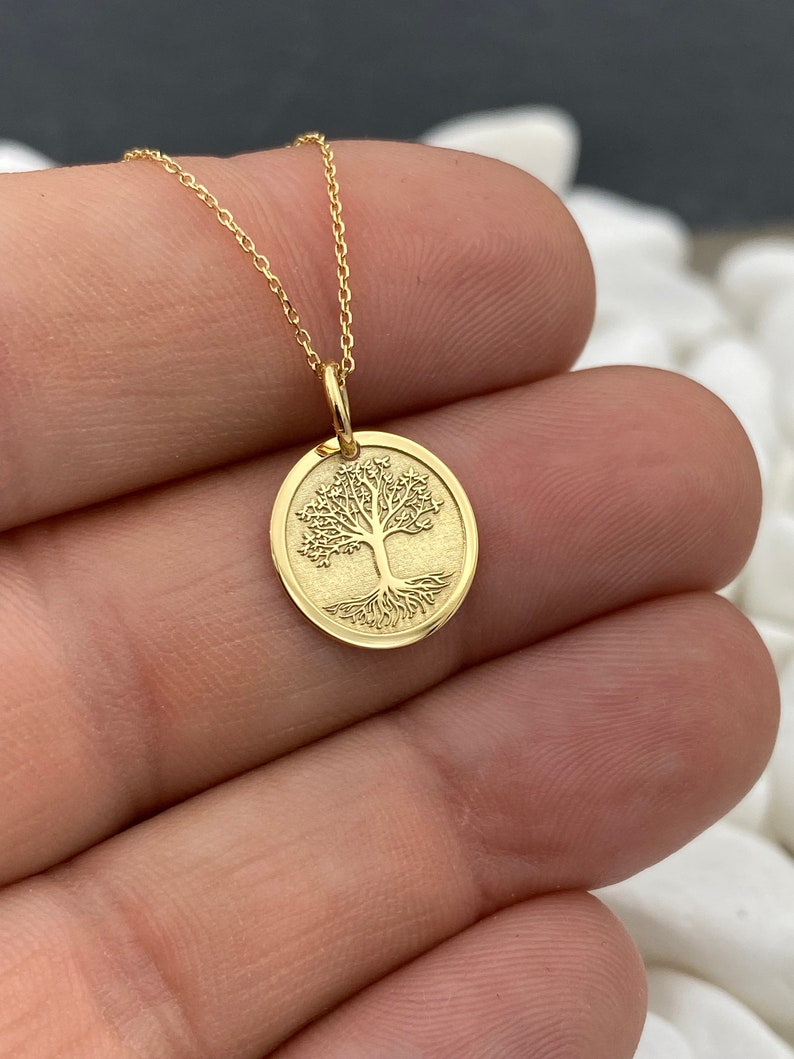May include: A gold-colored necklace featuring a circular pendant with a detailed tree design. The tree has a full crown and visible roots. The pendant hangs from a delicate gold chain, creating an elegant and symbolic piece of jewelry.
