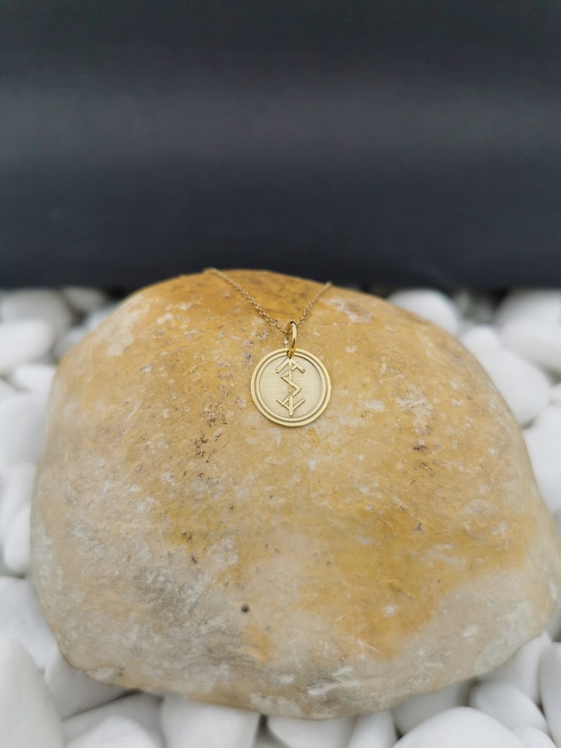 May include: A gold chain necklace with a round pendant featuring a stylized symbol. The pendant is on a light brown rock.