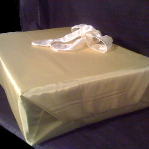 May include: A rectangular gift box wrapped in smooth, light gold paper. The top of the package is adorned with a large, decorative white satin ribbon bow. The wrapping is neatly folded at the corners, creating a clean and uniform appearance.