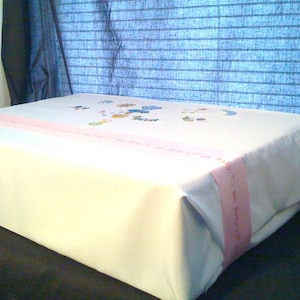 May include: A rectangular box wrapped in plain white paper, featuring a light pink ribbon band across the top. The top surface is decorated with small, colorful embroidered motifs of various shapes, including stars, a crescent moon, and small animals.