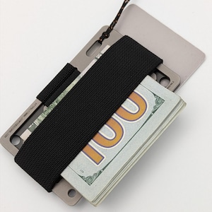 Puede incluir: Cartera metálica minimalista con una banda elástica negra que sujeta un fajo de billetes de 100 dólares estadounidenses. La cartera tiene un pequeño lazo para una correa. El texto "HTTP://WWW.JIMMYVOEDISONS.COM" es visible.