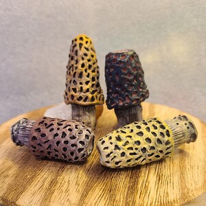 May include: Four ceramic mushroom sculptures in various colors and patterns. The mushrooms are arranged on a wooden surface.