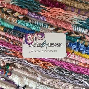 May include: A collection of colorful, handmade loc extensions in various shades of pink, blue, green, and neutral tones. The extensions are intricately wrapped and braided. A business card with the text "LOCKS BY LEVAN, LOCTICIAN & ACCESSORIES" is in the center.