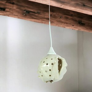 Puede incluir: Una lámpara colgante blanca con una pantalla esférica texturizada con múltiples agujeros. La luz cuelga de un cable blanco y está suspendida de un techo con vigas de madera.