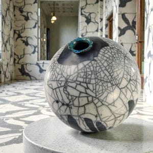 Puede incluir: Un jarrón esférico de cerámica con un esmalte blanco craquelado y detalles en negro. El borde superior es azul oscuro. El jarrón está sobre una mesa redonda gris moteada. El fondo presenta un pasillo con paredes y suelo estampados.