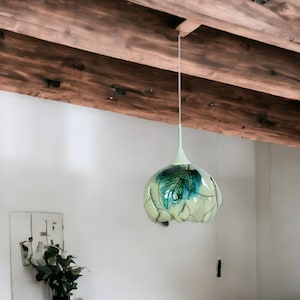 Puede incluir: Una lámpara colgante de vidrio blanco con un diseño de hoja verde y azul cuelga de una viga de madera del techo.