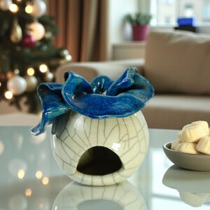 May include: A decorative ceramic bowl with a blue, flower-like top sits on a glass table. The bowl is white with a crackle pattern. A small bowl of white candies is to the right. A Christmas tree is in the background.