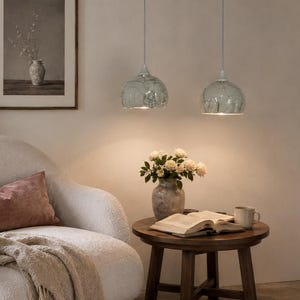 May include: Two light green pendant lights with a textured, dimpled surface hang from the ceiling above a round wooden side table. The table holds a ceramic vase with white flowers, an open book, and a mug, positioned next to a white armchair.
