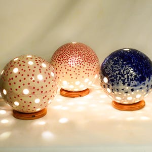 Op de afbeelding: Drie keramische globe lampen met verschillende patronen. De lampen zijn wit met rode, roze en blauwe stippen. De lampen zijn aan en geven een warm licht.