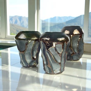 May include: Three ceramic vases with a distinctive, textured design. The vases have a dark, glossy finish with shades of blue, grey, and brown. The background features a scenic view of mountains and a bright, sunny sky.