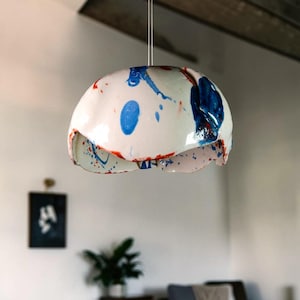 May include: A white ceramic pendant light with blue and red paint splatters. The light fixture is suspended from the ceiling and has a rounded shape with a scalloped edge. The interior is visible.