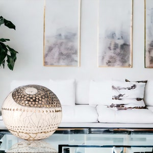 Op de afbeelding: Een witte en gouden decoratieve bol met een textuur zit op een glazen salontafel voor een witte bank met witte en bruine kussens met patronen. Drie ingelijste abstracte schilderijen hangen aan de muur achter de bank.