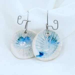 May include: A pair of white ceramic earrings with blue accents. The earrings are round and have a textured surface. They are attached to silver hooks.