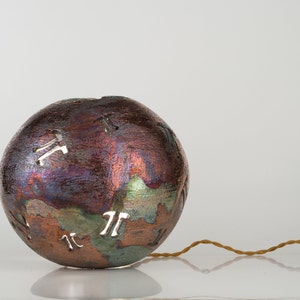 May include: A round, metallic, iridescent sphere with a textured surface and cutouts in the shape of the mathematical symbol pi. The sphere is attached to a thin, brown cord.