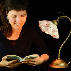 Może przedstawiać: Kobieta czyta książkę w ciepłym świetle lampy biurkowej w stylu vintage z białym i różowym abażurem w kwiaty.