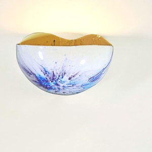 May include: A wall-mounted light fixture with a half-bowl shape. The fixture has a white interior with blue and purple splatter paint design. The top portion is a golden-brown color. The light is on, illuminating the wall.