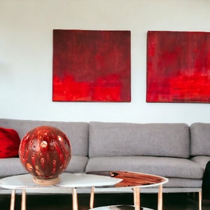 May include: A red ceramic sphere lamp with a textured surface sits on a coffee table in front of a gray sofa. Two red abstract paintings hang on the wall behind the sofa.