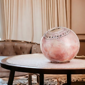 May include: A pink and white ceramic sphere with a decorative pattern sits on a round wooden table with a white surface. The table is in front of a white sofa with a tufted back.