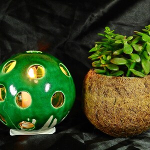 May include: A green ceramic candle holder with holes, shaped like a sphere, with a lit candle inside. The candle holder is sitting on a black surface next to a potted succulent plant in a coconut shell.