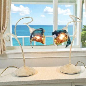 May include: Two decorative table lamps with a floral design. The lamps have a cream-colored base and stem, with a curved top supporting a blue and gold glass shade. The lamps are placed on a white countertop in front of a window.