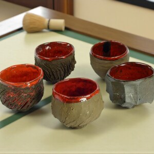 May include: Five handcrafted ceramic tea bowls with textured exteriors and vibrant red interiors. The bowls vary in shape and texture, showcasing a rustic aesthetic. A bamboo whisk rests in the background, suggesting a tea ceremony.