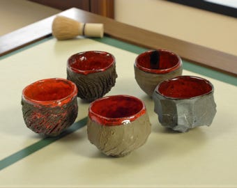 May include: Five handcrafted ceramic tea bowls with textured exteriors and vibrant red interiors. The bowls vary in shape and texture, showcasing a rustic aesthetic. A bamboo whisk rests in the background, suggesting a tea ceremony.