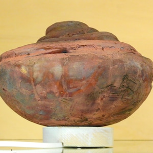 May include: A reddish-brown ceramic pot with a rounded shape and a raised, coiled lid. The pot has a rough, textured surface and appears to be ancient.