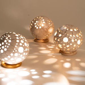 May include: Three white ceramic sphere lamps with cut-out designs. The lamps are lit and casting shadows on the white surface.