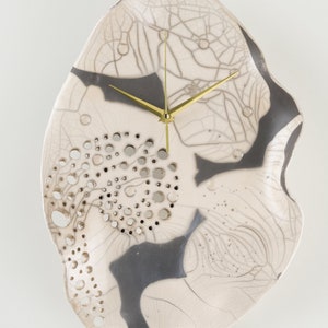 May include: A white ceramic clock with a grey and black design. The clock has a unique, organic shape and gold-coloured hour and minute hands. The clock face has a pattern of small holes.