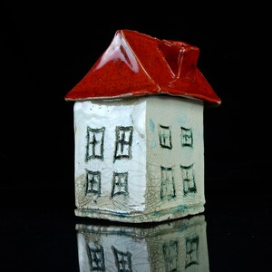 Könnte beinhalten: Ein kleines weißes Keramikhaus mit rotem Dach und blauen Fenstern. Das Haus hat eine strukturierte Oberfläche und steht auf einem schwarzen Hintergrund.