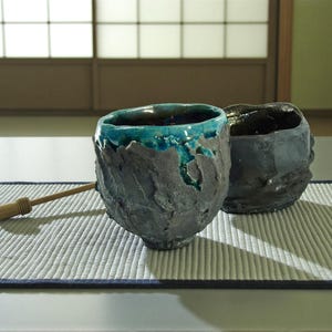May include: Two handcrafted ceramic tea bowls with textured, dark gray exteriors. One bowl has a turquoise interior and rim, the other a dark interior. A bamboo tea whisk rests on a woven mat, suggesting a tea ceremony.