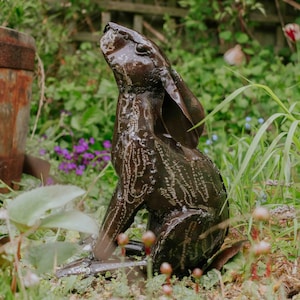 May include: A bronze metal hare sculpture, seated and looking upwards. The sculpture has a textured surface and is set in a garden with green plants and purple flowers. The hare is a popular garden ornament.
