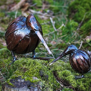 May include: Two metal sculptures of kiwi birds, one larger than the other, are perched on a bed of moss. The birds are made of recycled metal and have a rustic brown finish.